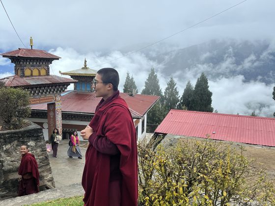 A person in a maroon robe stands on a grassy hill with misty mountains and traditional buildings in the background.