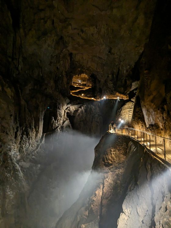 A dimly lit cave with a narrow walkway, illuminated by lights, winding through rocky formations and mist in the background.