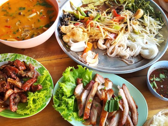 A table with a variety of Thai dishes, including a spicy soup, salad with noodles, grilled meats, and dipping sauce, served with fresh vegetables.