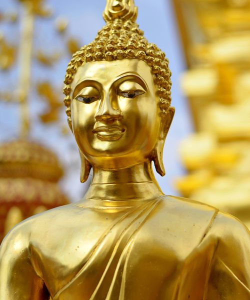 Golden Buddha statue with intricate details, set against a blurred temple background, under a clear blue sky.