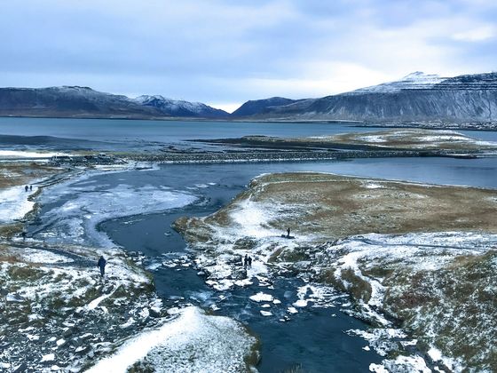 A snowy Icelandic landscape with icy water, distant mountains, and scattered people walking on paths under a cloudy sky.