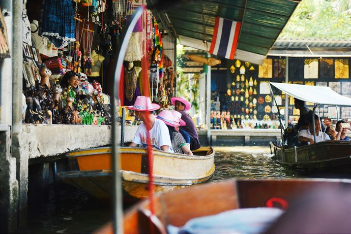 Boats navigate a bustling floating market with people in pink hats, surrounded by colorful souvenirs and a Thai flag overhead.