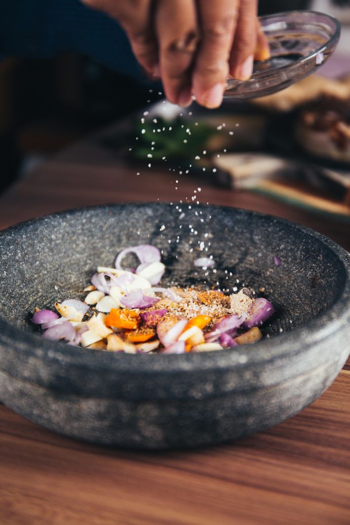 A hand sprinkles salt into a stone bowl containing sliced shallots, garlic, and turmeric on a wooden surface.