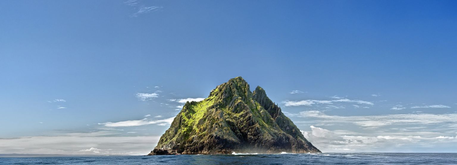 Rocky, green island with steep cliffs rises from a calm blue sea, under a clear sky with few clouds.