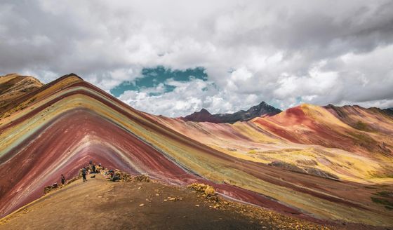 Colorful mountain landscape with layered red, yellow, and green hues under a cloudy sky, with people standing on a ridge.