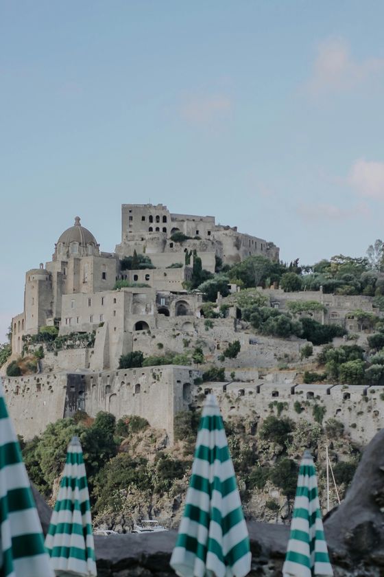 Castle on a hill with stone walls and domed tower, surrounded by greenery; striped umbrellas in the foreground.