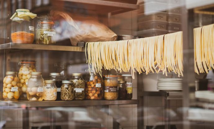 Jars of preserved foods and a row of fresh pasta hang in a kitchen with shelves, reflecting a cozy, rustic culinary setting.