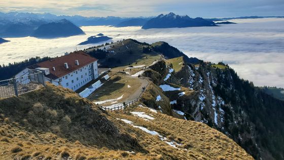 Mountain landscape with a building on a ridge, snow patches, and a sea of clouds below, framed by distant peaks under a blue sky.