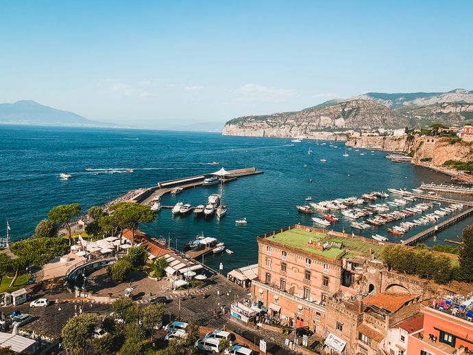 Coastal view of a marina with boats, buildings, and cliffs under a clear blue sky. Mountains are visible in the background.