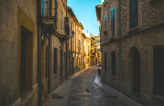 Narrow cobblestone street lined with rustic buildings and balconies under a clear blue sky.