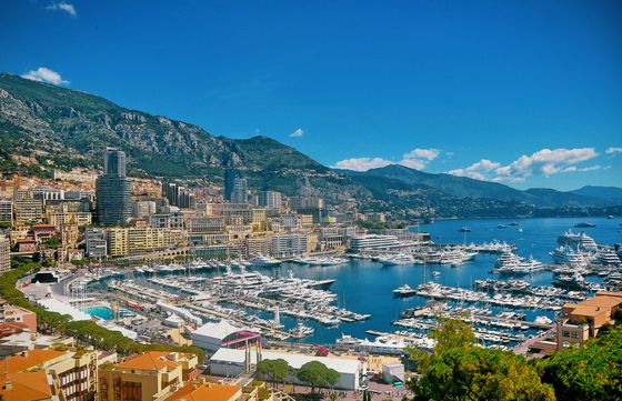 A coastal cityscape with numerous yachts in a marina, surrounded by mountains and buildings under a clear blue sky.