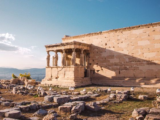 Ancient temple with Caryatid columns under a clear sky, surrounded by ruins and distant hills, bathed in warm sunlight.