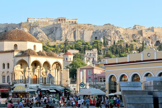 Bustling square in Athens with people, shops, and historic buildings. The Acropolis looms in the background under a clear blue sky.