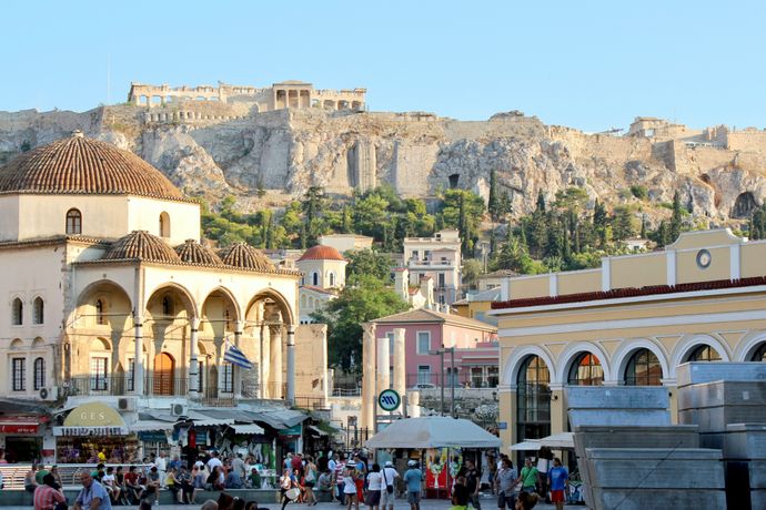 Bustling square in Athens with people, shops, and historic buildings. The Acropolis looms in the background under a clear blue sky.
