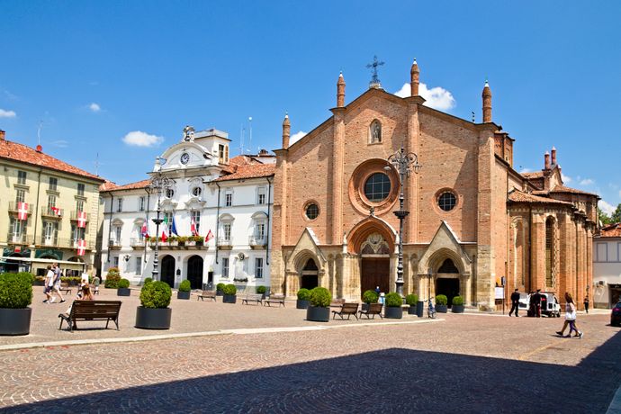 Historic brick church with a large rose window in an Italian square, surrounded by buildings and people walking on a sunny day.