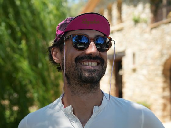 A smiling man wearing sunglasses and a cap stands outdoors on a sunny day, with a stone building and green foliage in the background.