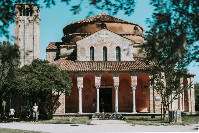 Historic brick church with a tall bell tower, arched entrance, and red-tiled roof, surrounded by trees under a clear blue sky.