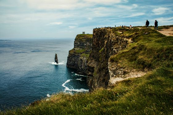 Cliffs of Moher with people walking along the edge, overlooking the ocean and a sea stack under a cloudy sky.