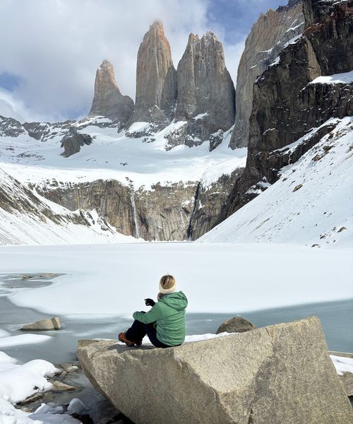 A person in a green jacket sits on a rock overlooking a snowy landscape with towering mountains and a frozen lake under a partly cloudy sky.