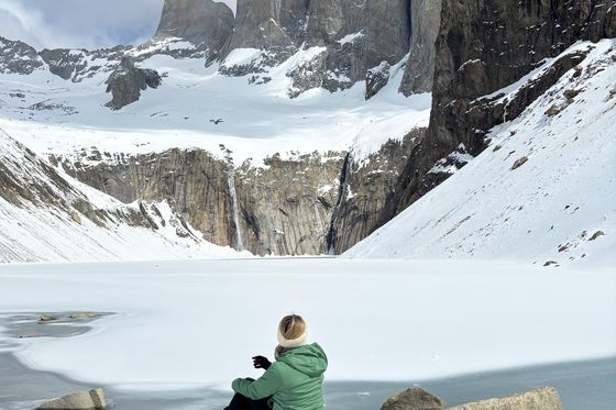 A person in a green jacket sits on a rock overlooking a snowy landscape with towering mountains and a frozen lake under a partly cloudy sky.