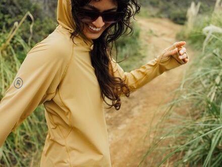 A woman wearing sunglasses and a yellow hoodie smiles while walking on a dirt path surrounded by greenery.