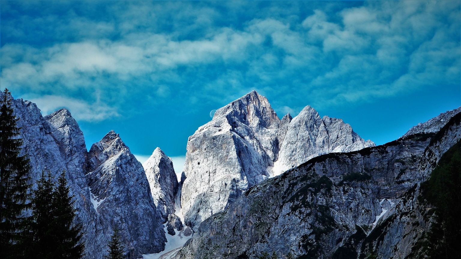 Snow-capped mountain peaks under a bright blue sky with scattered clouds, framed by dark evergreen trees.