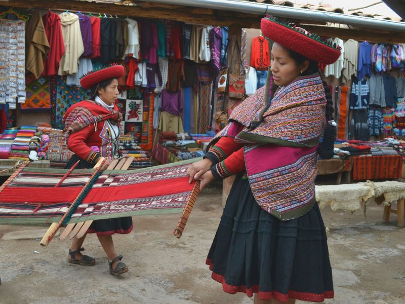 Two women in traditional attire weave on looms in a colorful market filled with textiles and garments.