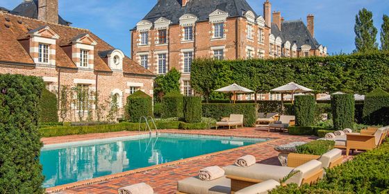 Luxurious estate with a large pool, surrounded by elegant brick buildings, manicured hedges, and cushioned lounge chairs under umbrellas.