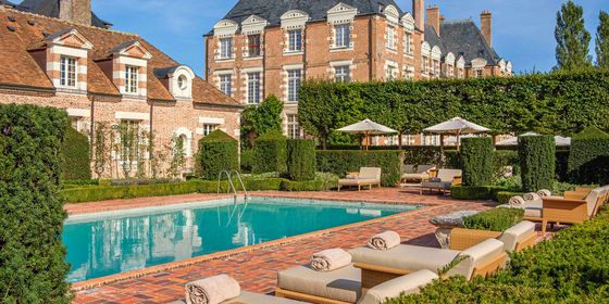Luxurious outdoor pool area with sun loungers, surrounded by manicured hedges and historic brick buildings under a clear blue sky.