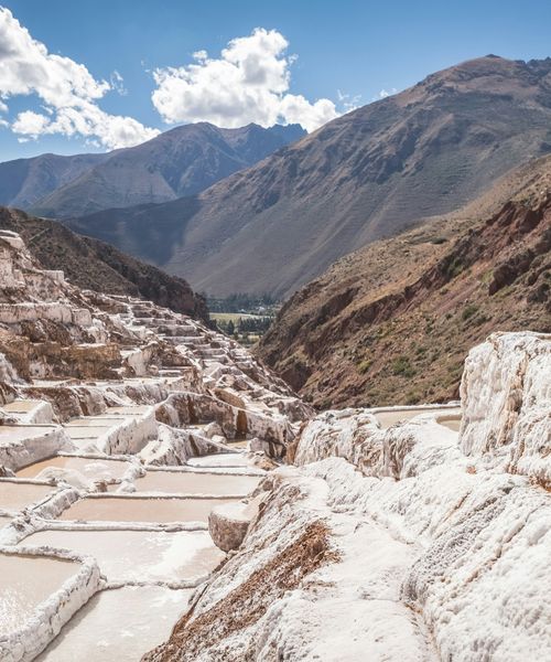 Terraced salt evaporation ponds on a mountainside with a valley and mountains in the background under a partly cloudy sky.