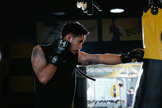 A person with tattoos punches a yellow boxing bag in a dimly lit gym, wearing black gloves and a sleeveless shirt.
