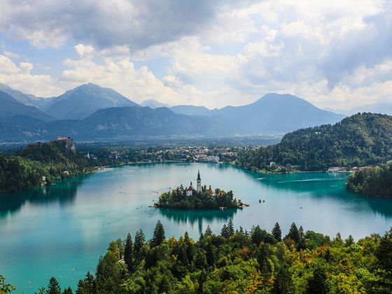 A scenic view of Lake Bled, with a small church on an island, surrounded by lush hills and distant mountains under a partly cloudy sky.