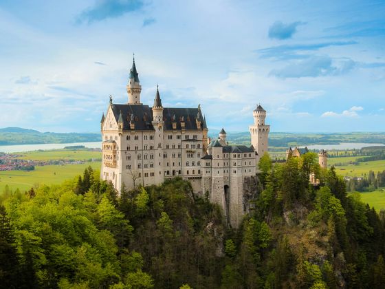 Neuschwanstein Castle perched on a lush hilltop, surrounded by forests and fields under a blue sky with scattered clouds.