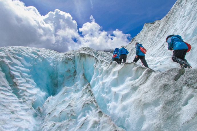 Three people in blue jackets climb a snowy glacier under a bright blue sky with clouds.