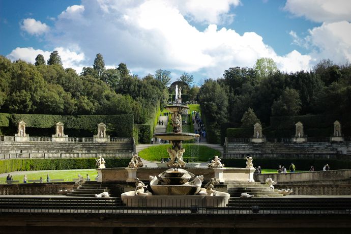 A grand tiered fountain with statues in a formal garden, surrounded by manicured hedges and trees under a partly cloudy sky.