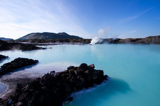 A serene geothermal lagoon with steaming water, surrounded by rocky terrain and distant mountains under a clear blue sky.