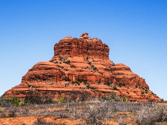 Red sandstone formation against a clear blue sky, resembling a layered mountain, surrounded by low vegetation and dry grassland.