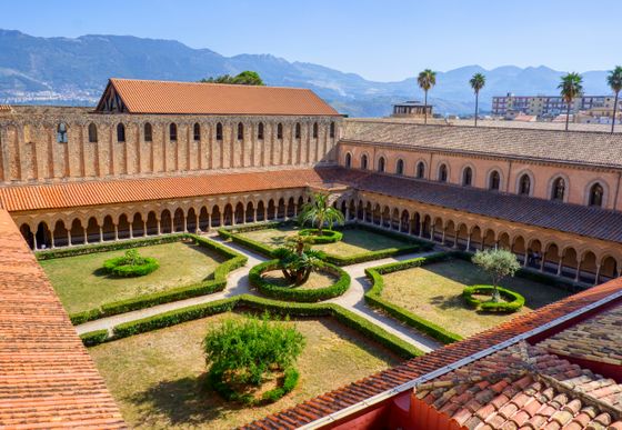 Aerial view of a historic cloister with a central garden, surrounded by arched walkways and red-tiled roofs, set against a mountainous backdrop.