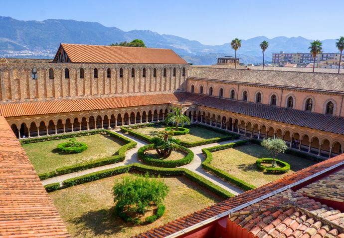 Aerial view of a historic cloister with a central garden, surrounded by arched walkways and red-tiled roofs, set against a mountainous backdrop.