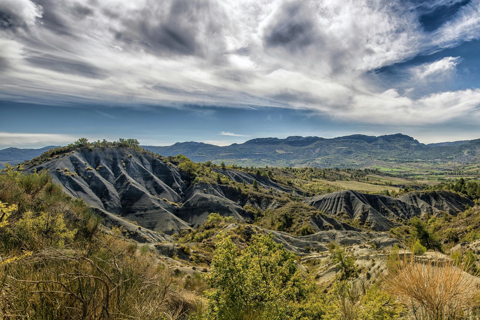 Rugged landscape with eroded hills, sparse vegetation, and a dramatic cloudy sky, set against distant mountains.