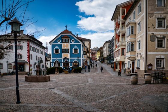 A charming European village square with colorful buildings, cobblestone streets, and a few people walking under a partly cloudy sky.