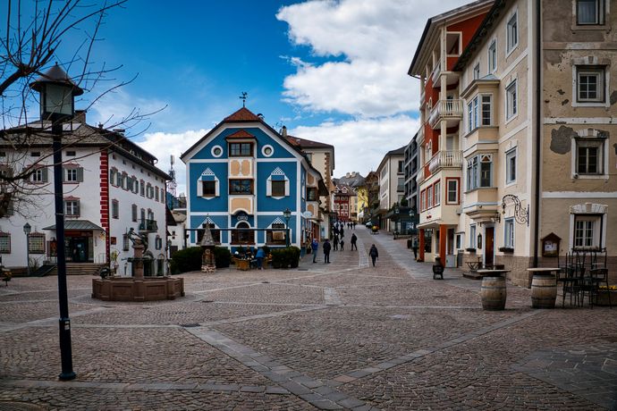 A charming European village square with colorful buildings, cobblestone streets, and a few people walking under a partly cloudy sky.