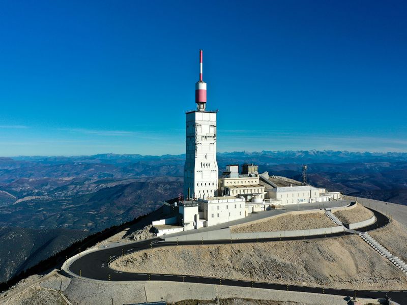 Aerial view of Mont Ventoux's summit with a white and red communication tower, surrounded by a winding road and mountainous landscape.