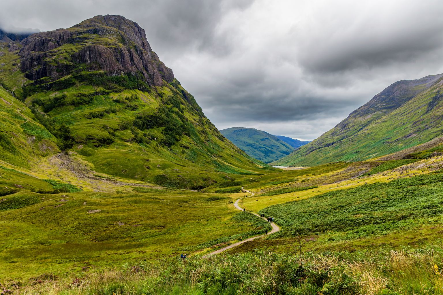 A winding path through a lush green valley flanked by steep hills under a cloudy sky.
