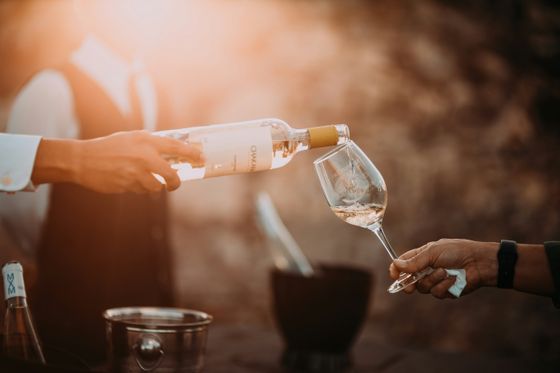 A person pours white wine from a bottle into a glass at an outdoor event, with warm sunlight in the background.