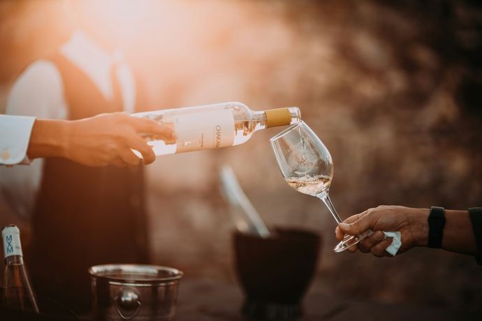 A person pours white wine from a bottle into a glass at an outdoor event, with warm sunlight in the background.