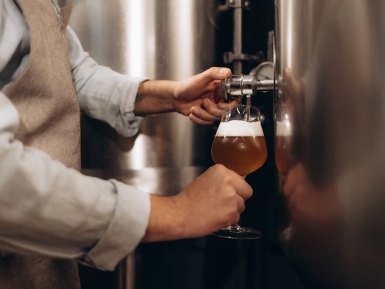 Person wearing an apron pours beer from a large metal brewing tank into a glass.
