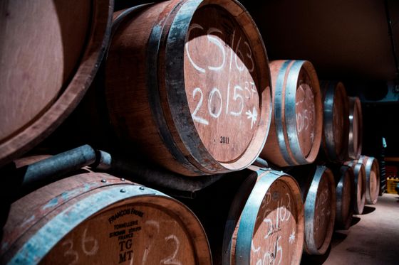 Stacked wooden wine barrels in a dimly lit cellar, each marked with white chalk inscriptions, including dates and codes.