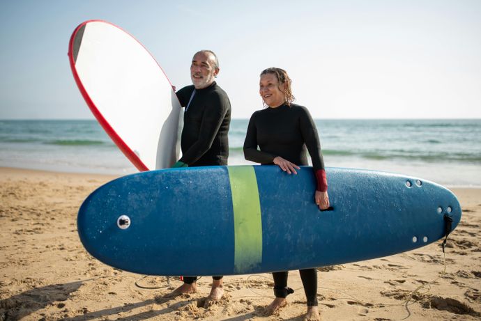 Two people in wetsuits holding surfboards stand on a sandy beach with the ocean in the background, smiling under a clear sky.
