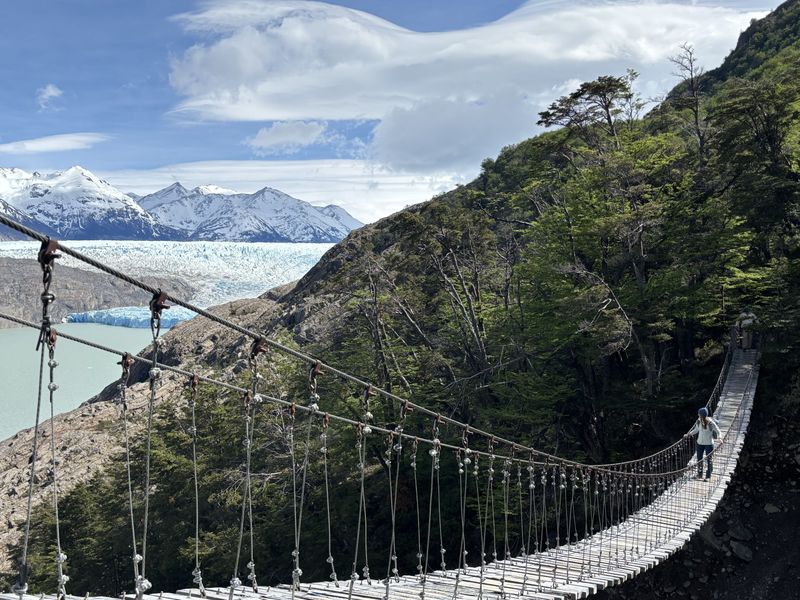 A person walks on a suspension bridge through a forest, with snow-capped mountains and a glacier in the background under a partly cloudy sky.